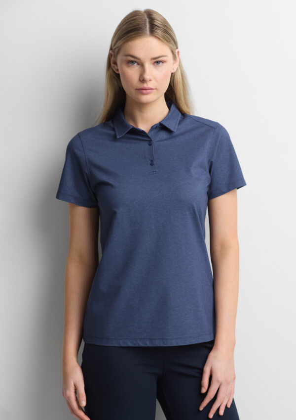 Woman wearing a blue polo shirt and navy pants, standing against a light gray wall.