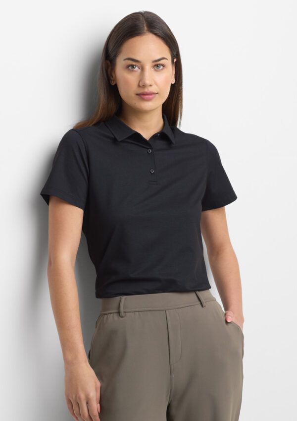 Woman standing against a white wall, wearing a black polo shirt and taupe trousers.