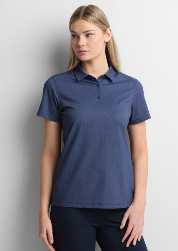 Woman wearing a blue polo shirt with short sleeves, standing against a light wall.