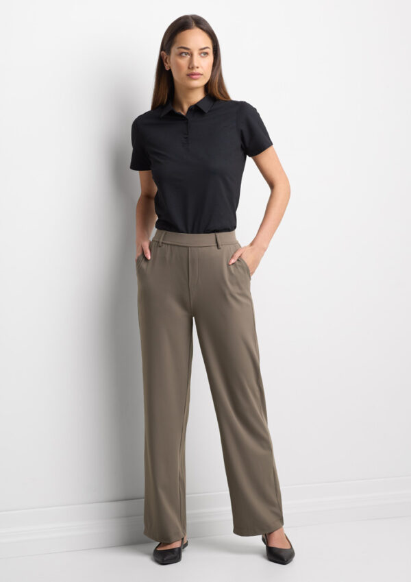 Full-length image of a woman standing against a white wall, wearing a black polo shirt and taupe wide-leg pants with hands in pockets.