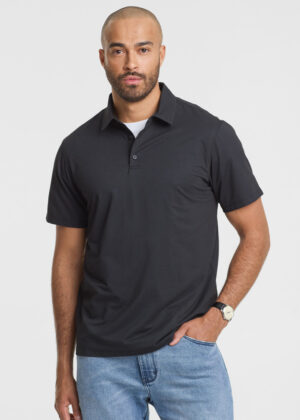 Man wearing a black polo shirt and light-wash jeans, standing with one hand in his pocket against a white background.