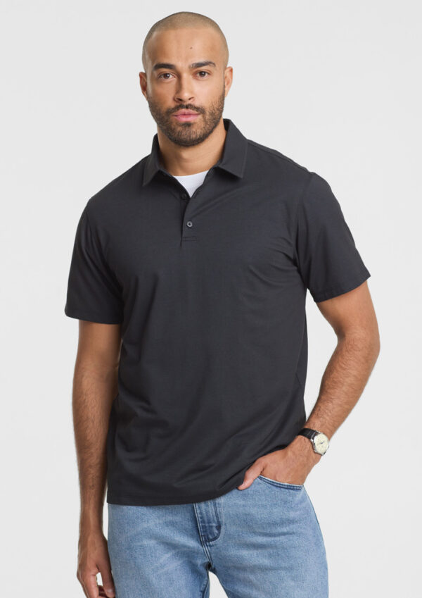 Man wearing a black polo shirt and light-wash jeans, standing with one hand in his pocket against a white background.
