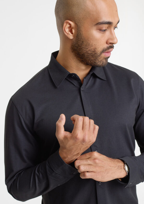 Man in a black button-down shirt adjusts his cuff, looking to the side.