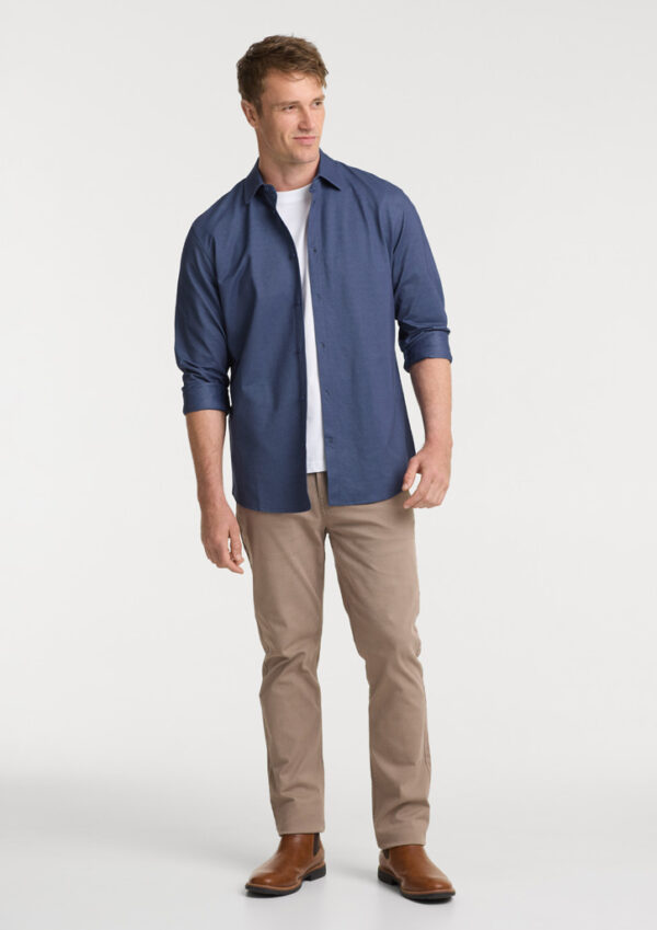 Man wearing a blue button-down shirt over a white T-shirt, beige chinos, and brown Chelsea boots in a full-body pose.