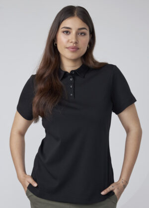 Woman posing in a black polo shirt with short sleeves, hands in pockets, against a light gray background.