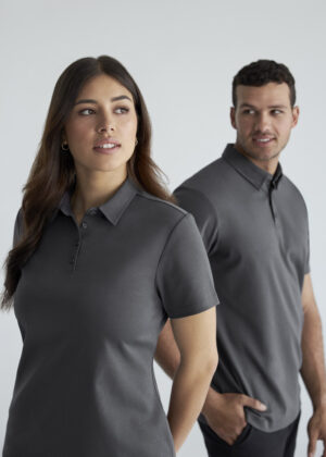 Woman in a dark gray polo shirt in the foreground with a man in a matching polo shirt behind her in a studio setting.