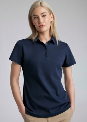 Woman with blonde hair wearing a navy polo shirt and beige pants, standing against a light gray background with hands in pockets.