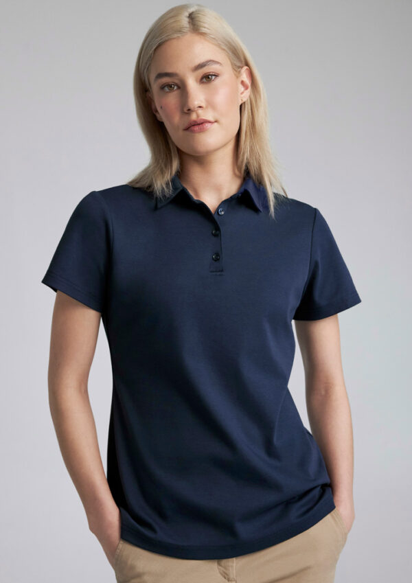 Woman with blonde hair wearing a navy polo shirt and beige pants, standing against a light gray background with hands in pockets.