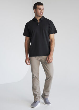 Man wearing a black polo shirt, beige pants, and gray sneakers standing in a studio setting.