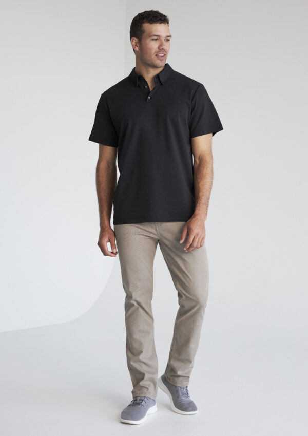 Man wearing a black polo shirt, beige pants, and gray sneakers standing in a studio setting.