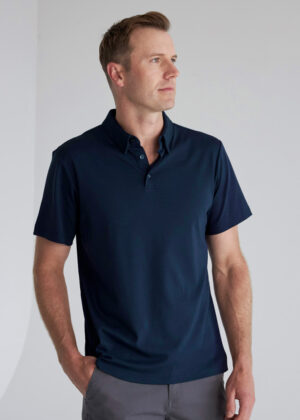 Man wearing a navy polo shirt and gray pants stands with hands in pockets, looking to the right against a light background.
