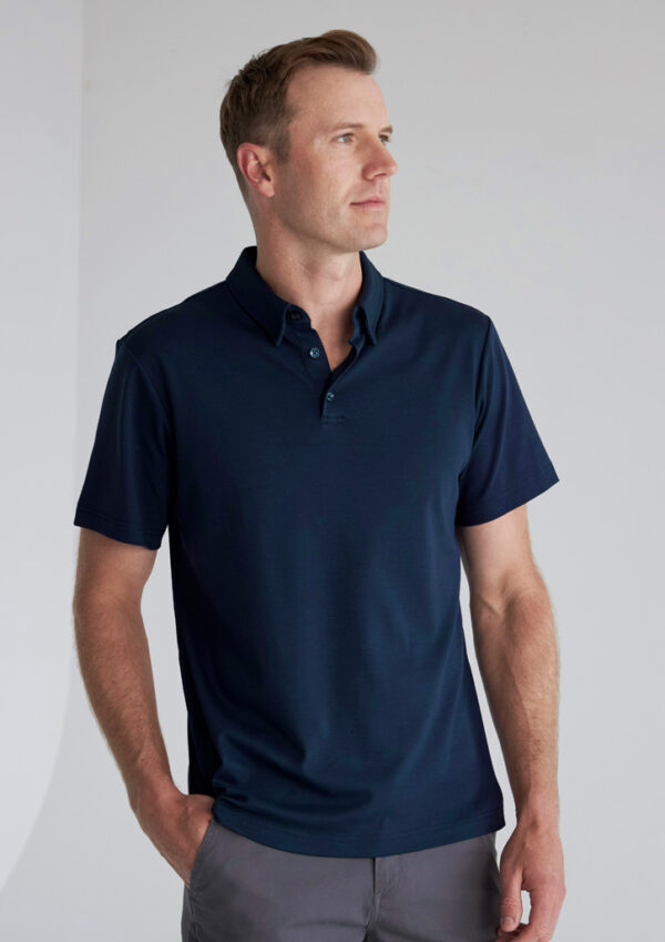Man wearing a navy polo shirt and gray pants stands with hands in pockets, looking to the right against a light background.