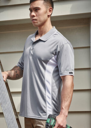 Man in a gray polo shirt with white side panels stands outdoors beside a ladder, holding a green cordless drill.