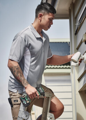 Man on a ladder installs an outdoor security camera on the side of a house with a cordless drill.
