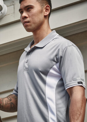 Man wearing a light grey polo shirt with white side panels, standing outdoors beside a building.