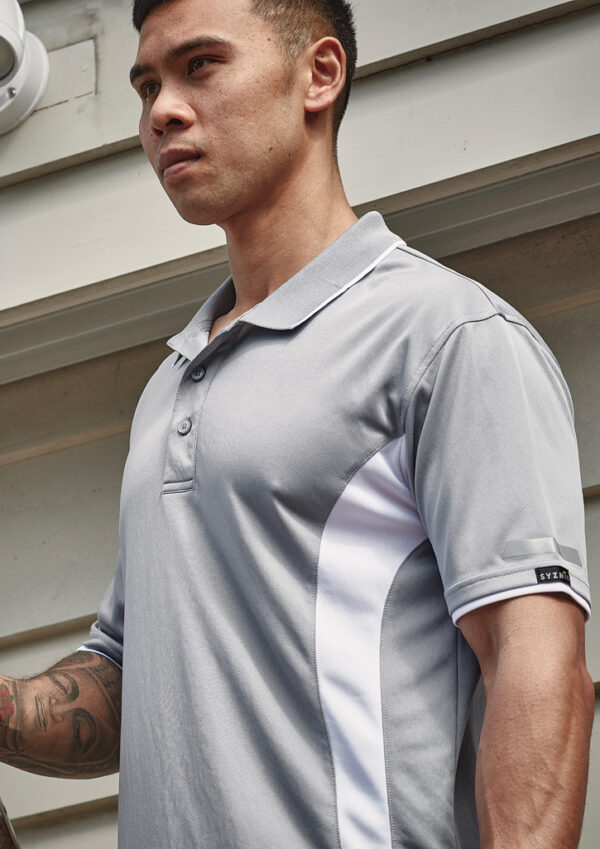 Man wearing a light grey polo shirt with white side panels, standing outdoors beside a building.