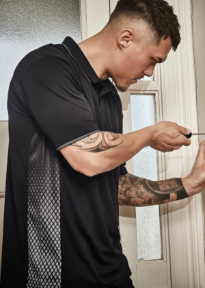 Man with tattoos in a black polo leaning by a doorway, looking at a handheld device (phone).