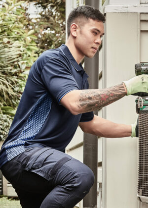 Man in a navy polo kneels beside an outdoor air conditioner, using a drill while wearing light green gloves.