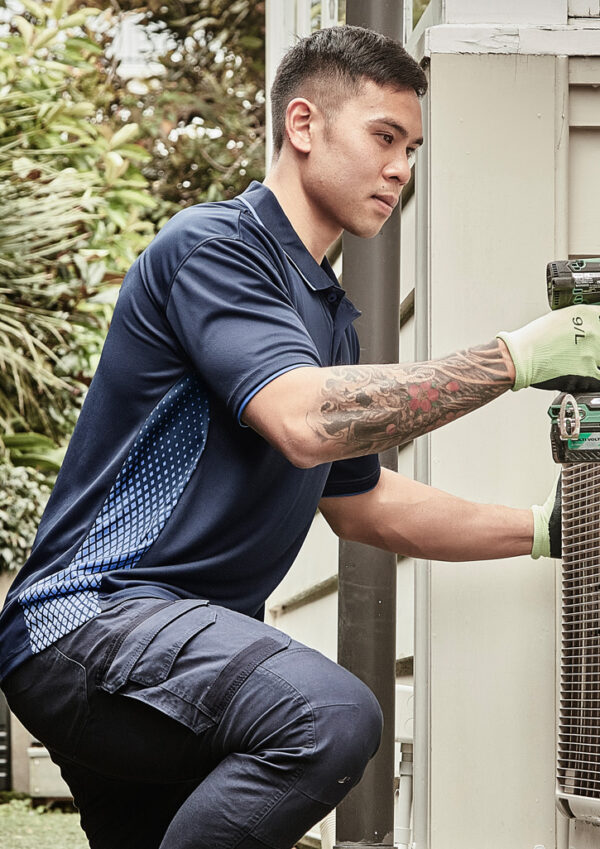 Man in a navy polo kneels beside an outdoor air conditioner, using a drill while wearing light green gloves.