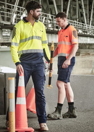 Two construction workers in high-visibility clothing stand near orange traffic cones, inspecting equipment.