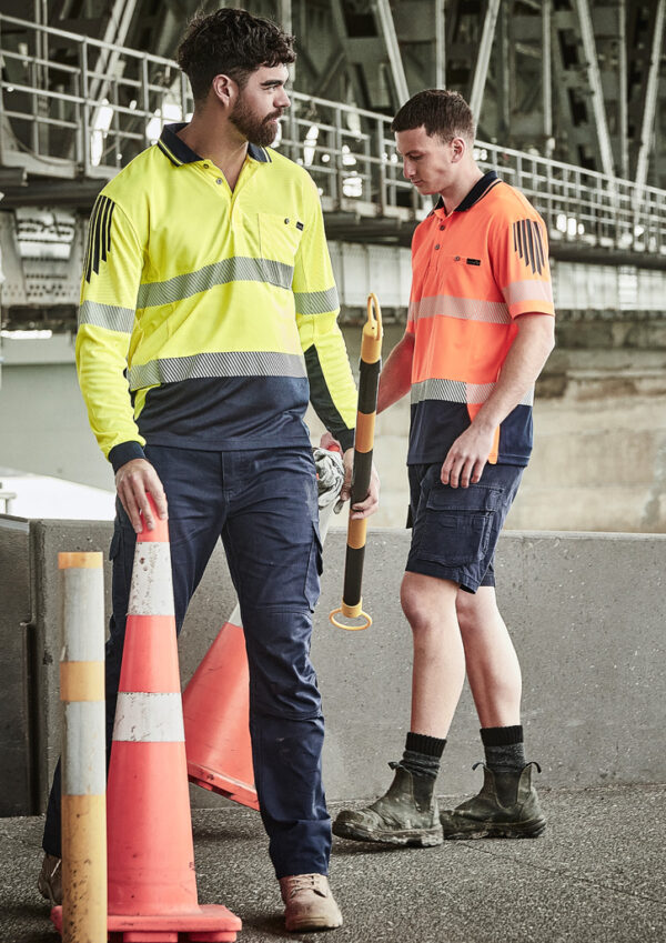 Two construction workers in high-visibility clothing stand near orange traffic cones, inspecting equipment.