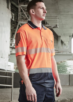 Man in a bright orange high-visibility polo with reflective stripes, standing under a concrete structure by the water.