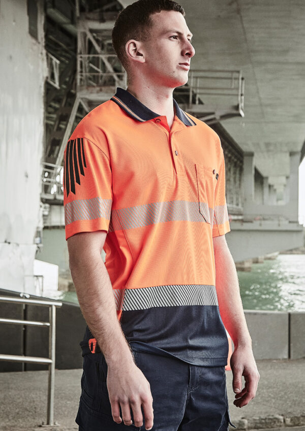 Man in a bright orange high-visibility polo with reflective stripes, standing under a concrete structure by the water.