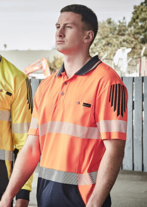 Man in an orange high-visibility polo shirt stands outdoors beside a fence, looking ahead, with another worker in yellow partially visible beside him.
