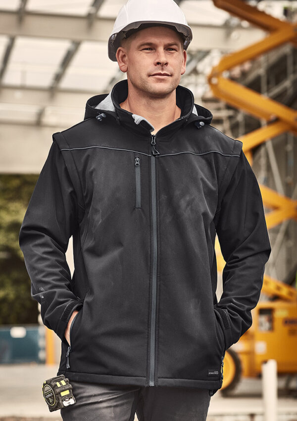 Construction worker in a black safety jacket and white hard hat, hands in pockets on a construction site with yellow equipment in the background.