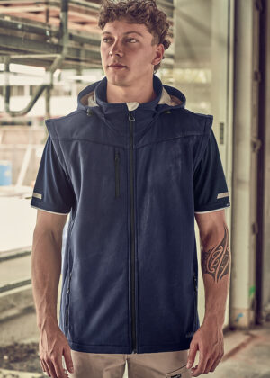 Young man in a navy sleeveless zip-up jacket, standing in an industrial setting; tattoo on his left forearm.