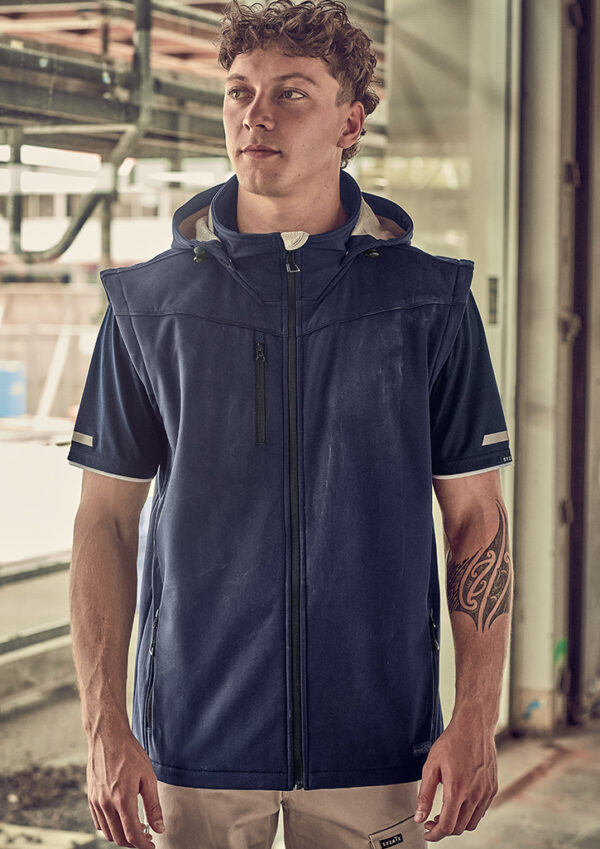 Young man in a navy sleeveless zip-up jacket, standing in an industrial setting; tattoo on his left forearm.