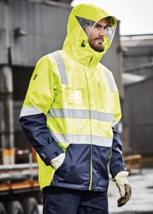 Man in a neon yellow high-visibility raincoat with reflective stripes and hood, standing outdoors at an industrial site.