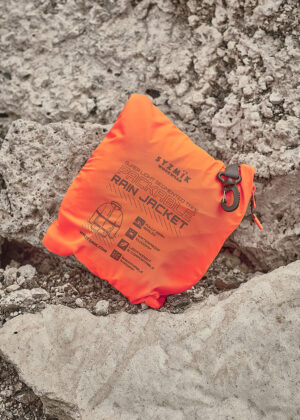 Bright orange packable rain jacket in its waterproof stuff sack on rough rocky terrain.