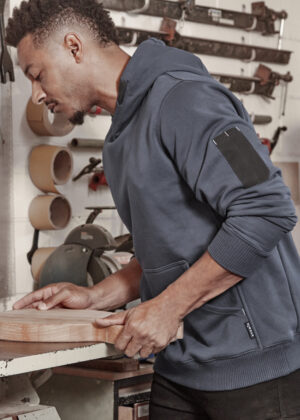 Man in a blue hoodie holding and guiding a wooden board on a workshop table with tools in the background.