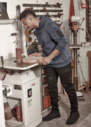 Man in a blue hoodie sanding a wooden board at a workshop bench, with woodworking tools and machines in the background