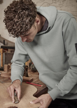 Young man in a light gray hoodie planes a wooden board at a workbench in a workshop.