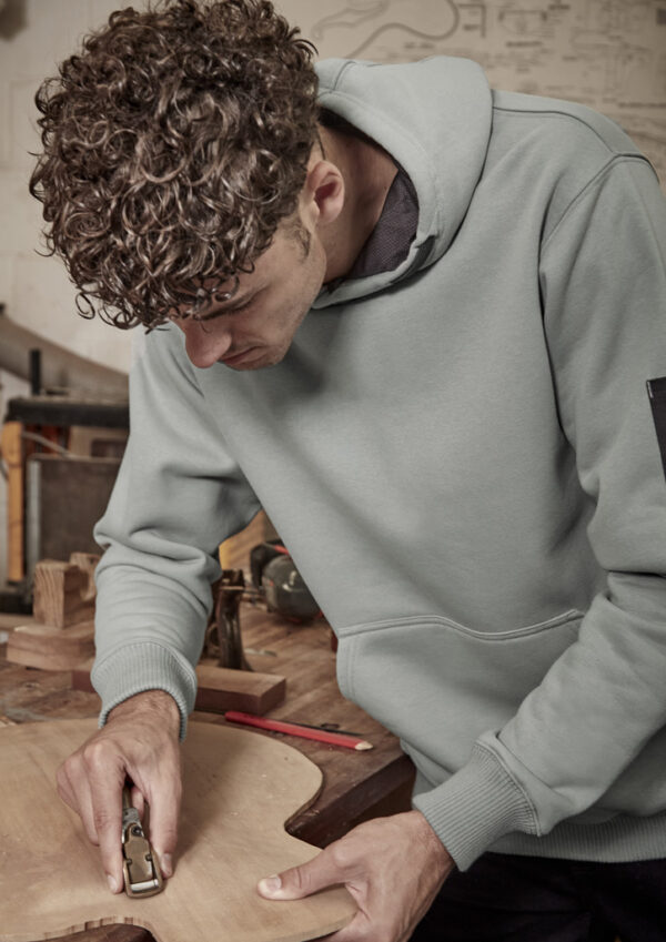 Young man in a light gray hoodie planes a wooden board at a workbench in a workshop.