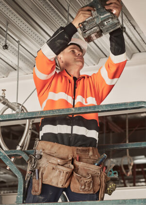 Construction worker in an orange safety jacket and white helmet using a drill to install ceiling panels. The tool belt is loaded with gear.
