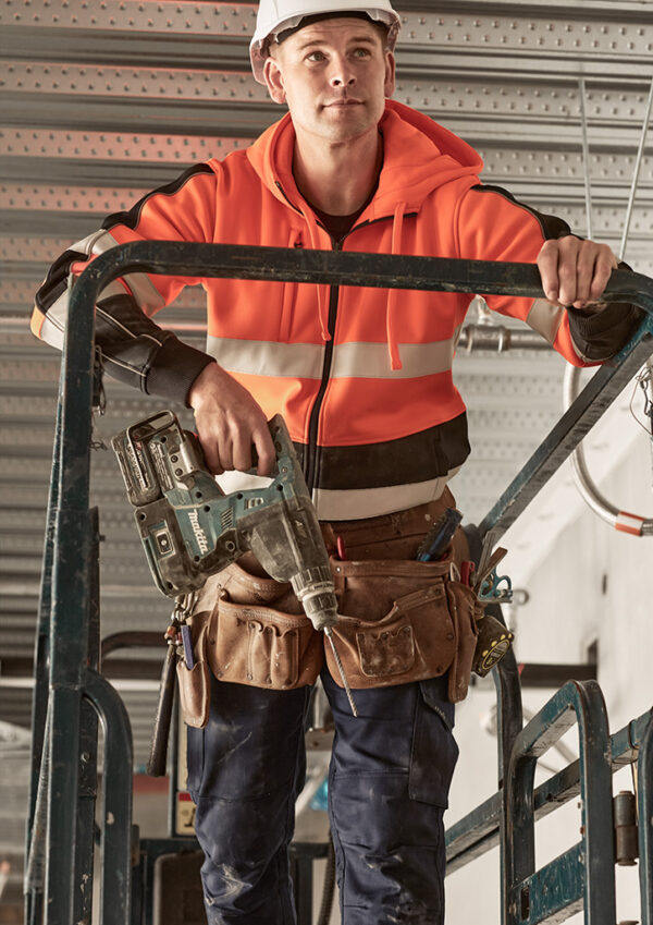 Construction worker in a bright orange safety jacket and hard hat, gripping a drill on a metal scaffold.