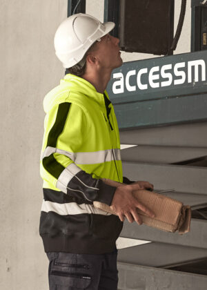 Construction worker in a neon safety jacket and white hard hat holding a cardboard box beside industrial equipment.