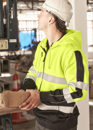 Construction worker in a high-visibility jacket and hard hat placing a cardboard box on a pallet in a warehouse or industrial setting.