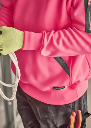 Close-up of a person in a bright pink jacket with a neon green glove gripping white cables in a work setting.