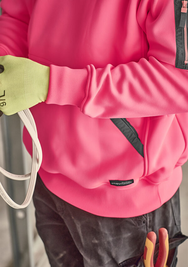 Close-up of a person in a bright pink jacket with a neon green glove gripping white cables in a work setting.