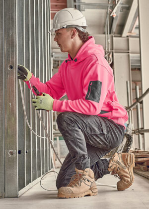 Construction worker in a pink hoodie and white hard hat kneels, installing or wiring cables into a metal wall frame.