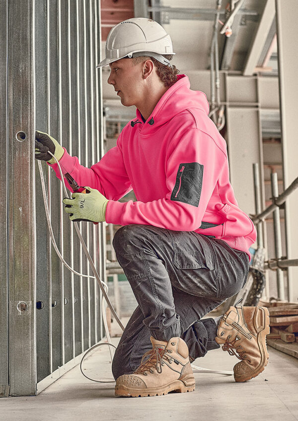 Construction worker in a pink hoodie and white hard hat kneels, installing or wiring cables into a metal wall frame.