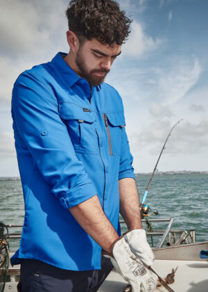 Bearded man in a blue shirt and gloves on a boat, handling a fish with the ocean behind him. (Informative: fishing scene, foreground subject handling catch)