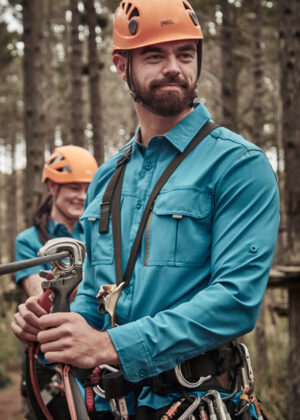 Man in teal shirt and orange helmet securing climbing rope in an outdoor forest setting; safety gear visible.