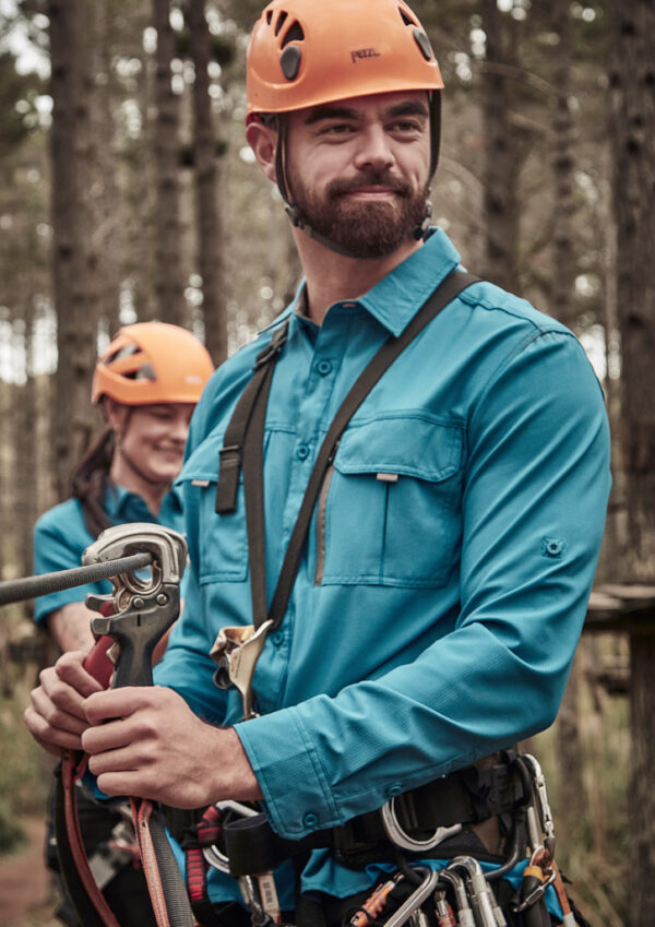 Man in teal shirt and orange helmet securing climbing rope in an outdoor forest setting; safety gear visible.