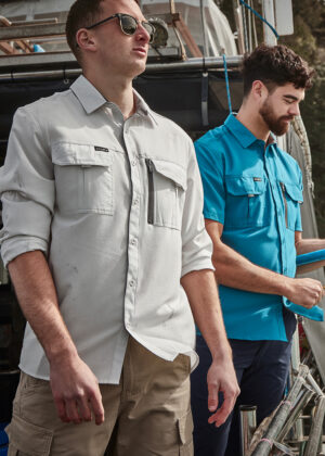 Two men in casual work shirts stand on a boat deck; the man in front wears sunglasses and beige shorts. Behind him, a second man in a blue shirt looks down.