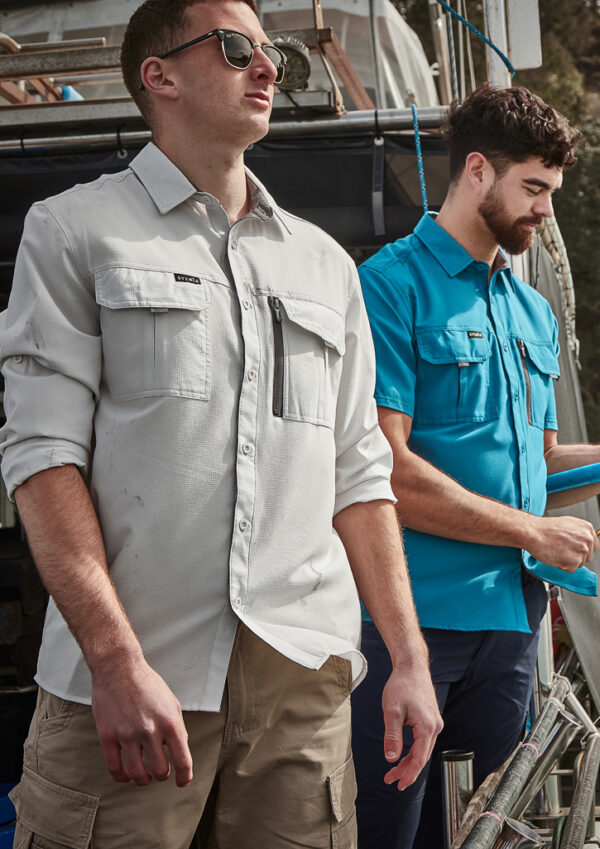 Two men in casual work shirts stand on a boat deck; the man in front wears sunglasses and beige shorts. Behind him, a second man in a blue shirt looks down.